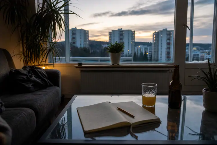 Olohuoneen pöydällä on muistikirja, mehulasillinen ja kaljapullo, ja ikkunasta näkyy Hervannan kerrostaloja.