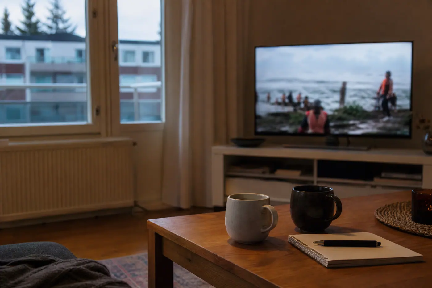 Hervantalaisen olohuoneen pöydällä on kaksi mukia ja muistikirja, taustalla televisio näyttää epätarkkaa kansainvälistä uutislähetystä.