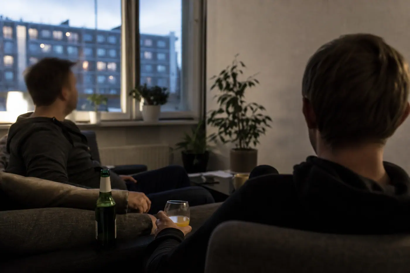 Hervantalaisen olohuoneen sohva, jolla istuu kaksi tunnistamatonta miestä; pöydällä on juomalasi ja pullo.