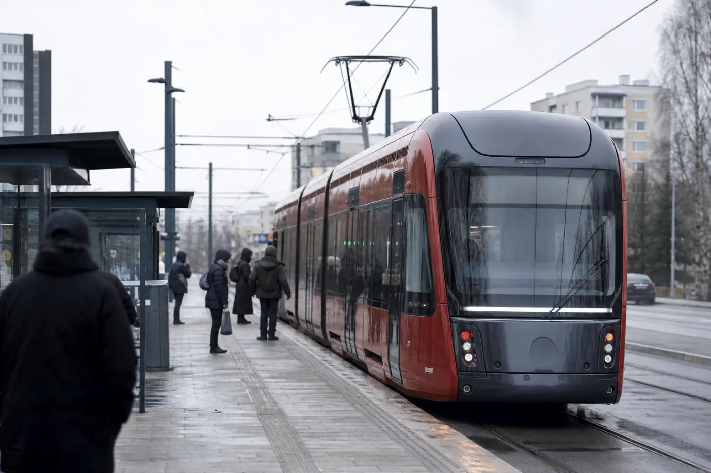 Ratikka pysähtyy Hervannassa harmaana päivänä, taustalla näkyy asuinrakennuksia ja muutamia epätarkkoja matkustajia.
