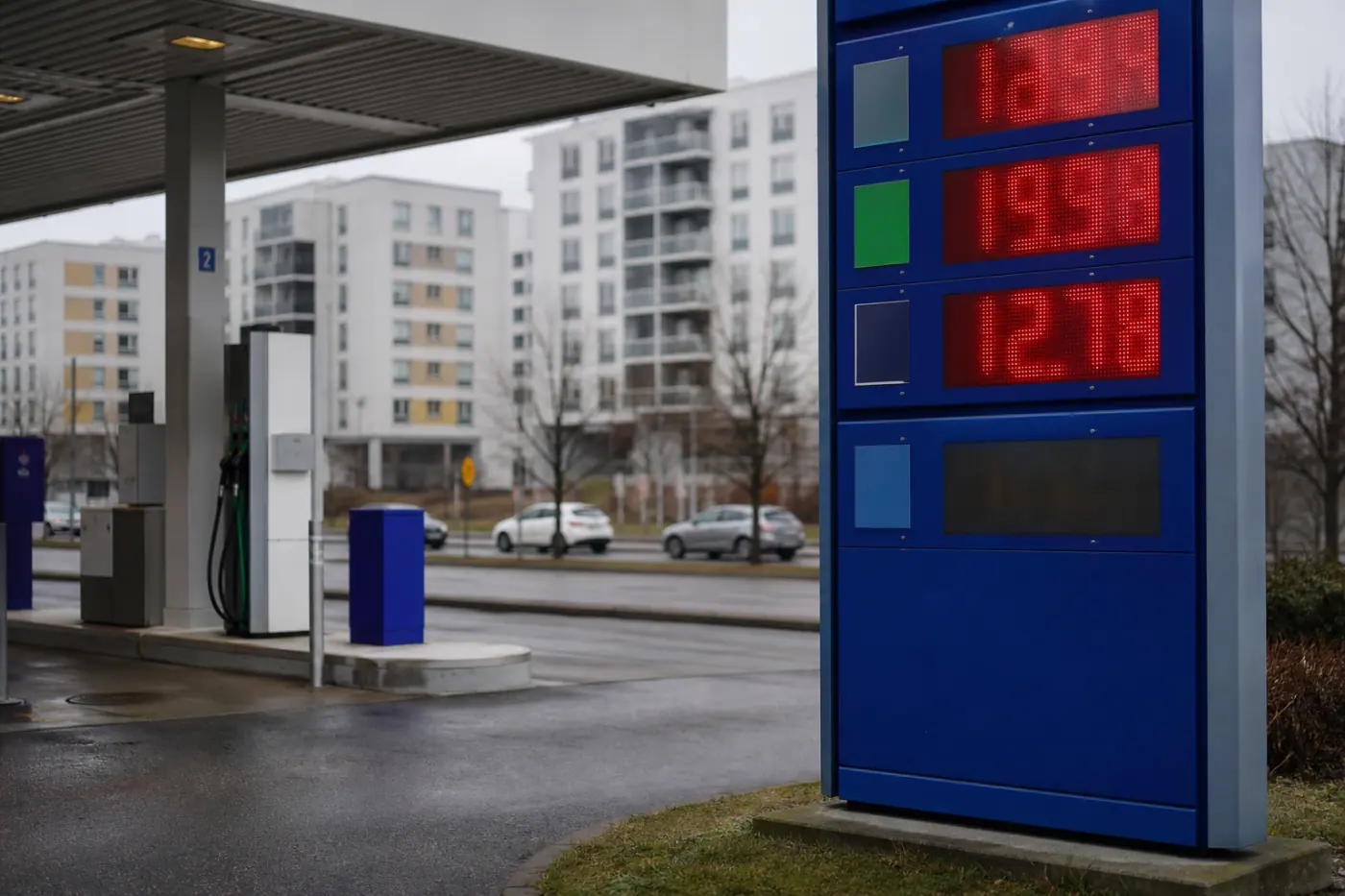 Polttoaineaseman mittareita ja hintataulu harmaassa kaupunkimaisemassa, taustalla autoja.
