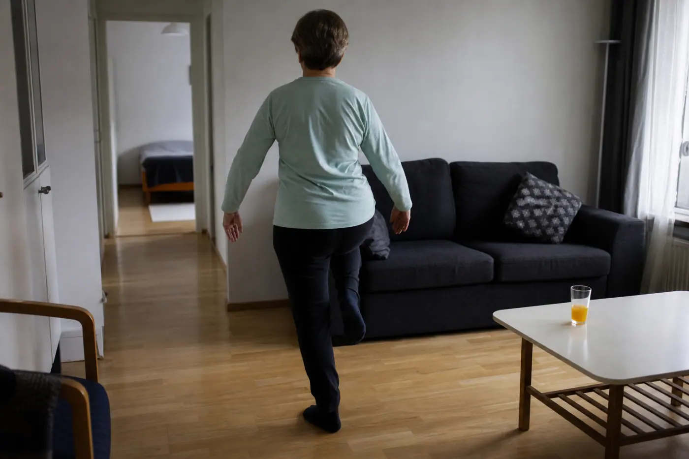 Olohuoneessa selin näkyvä henkilö tekee tasapainoharjoitusta sohvan edessä.