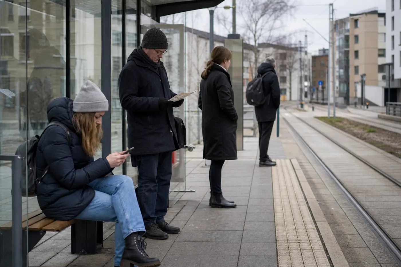 Suomalaisella joukkoliikenteen pysäkillä odottaa matkustajia; yksi katsoo puhelinta ja toinen pitää kädessään paperista aikataulua.