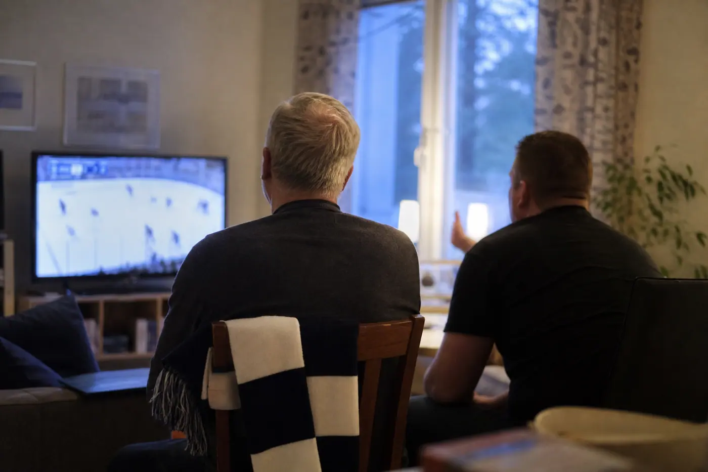 Hervantalaisen olohuoneen televisiossa näkyy epätarkka jääkiekkolähetys, ja kaksi selin kuvattua miestä keskustelee sen äärellä.